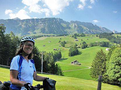 Foto2: Fit in den Radl-Sommer, Radreisen. Frau mit Fahrrad steht vor einem grünen Tal, im Hintergrund sind Berge zu sehen